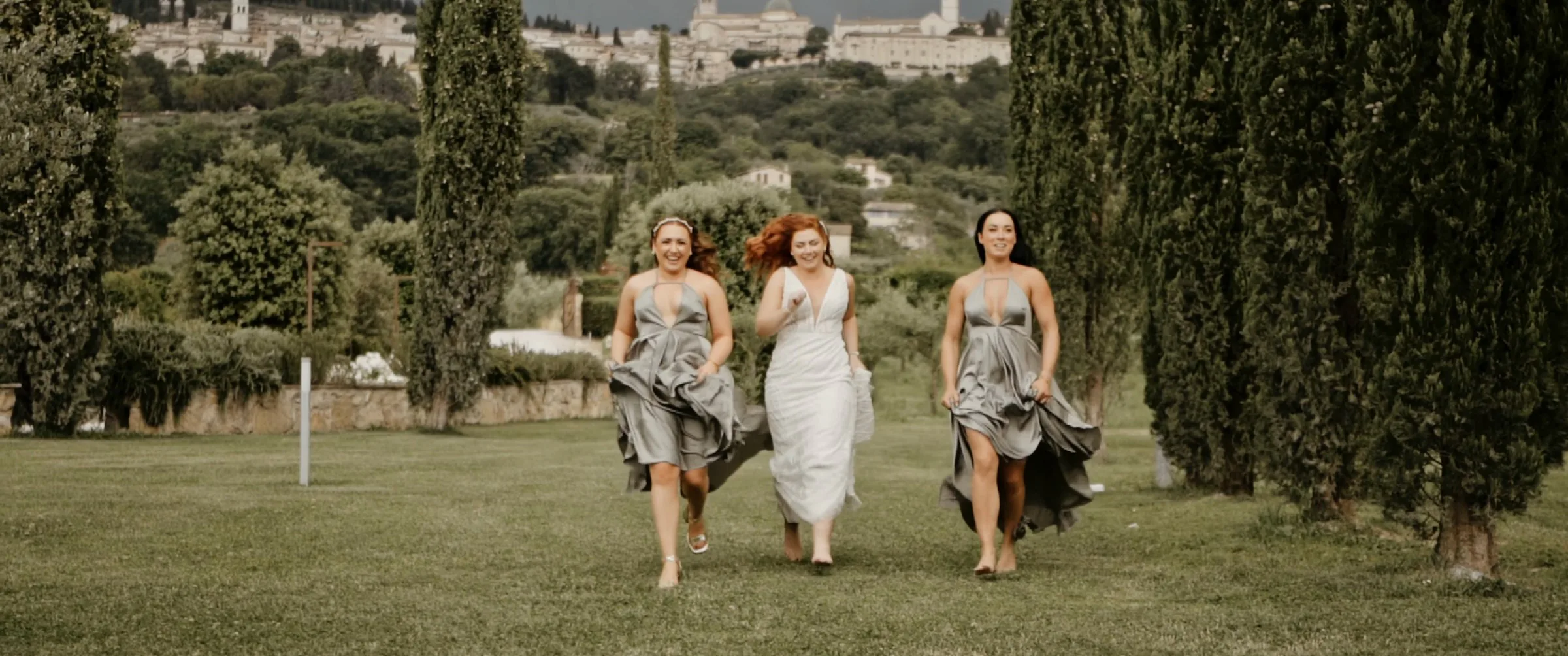 Bridesmaids running through the garden among the cypress trees at Tenuta di San Masseo, with Assisi in the background, a frame from James and Stacey’s film